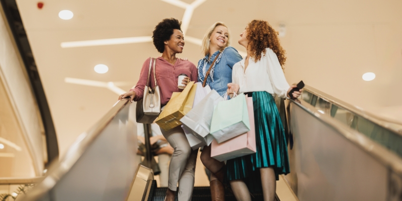 Three women stroll down an escalator with shopping bags, reflecting the bustling shopping experience on Las Olas Boulevard, Fort Lauderdale.